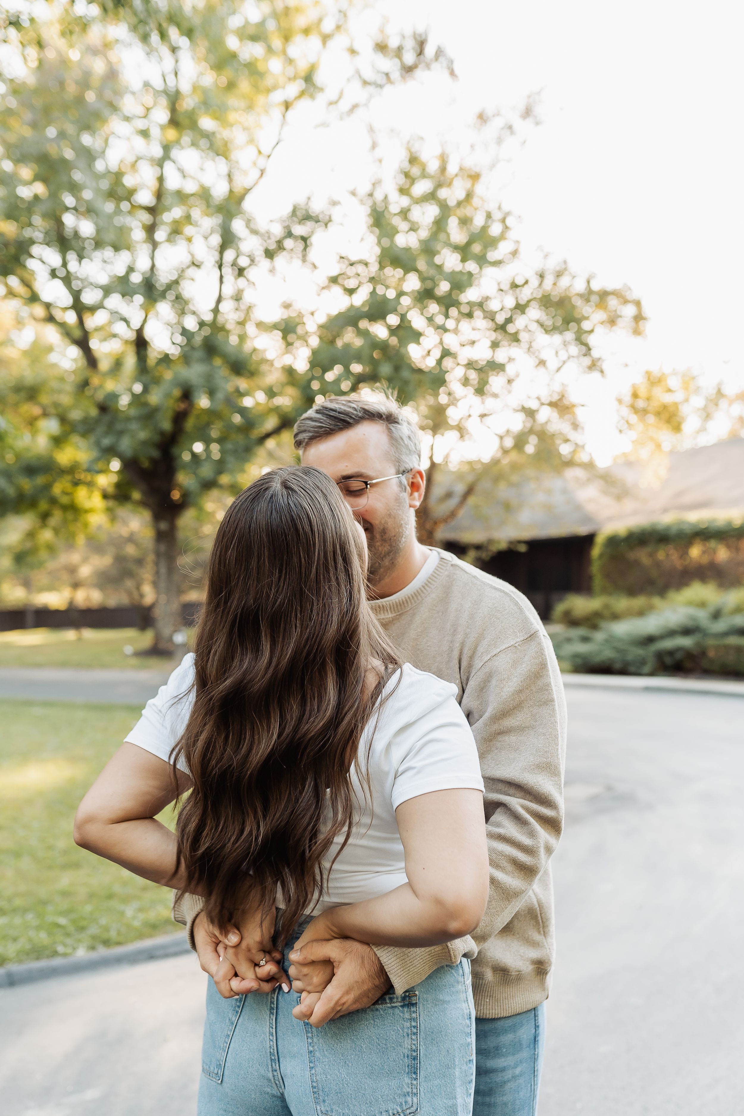 A couple shares a tender kiss outdoors in a sunlit park, with the woman's long dark hair flowing down her back.