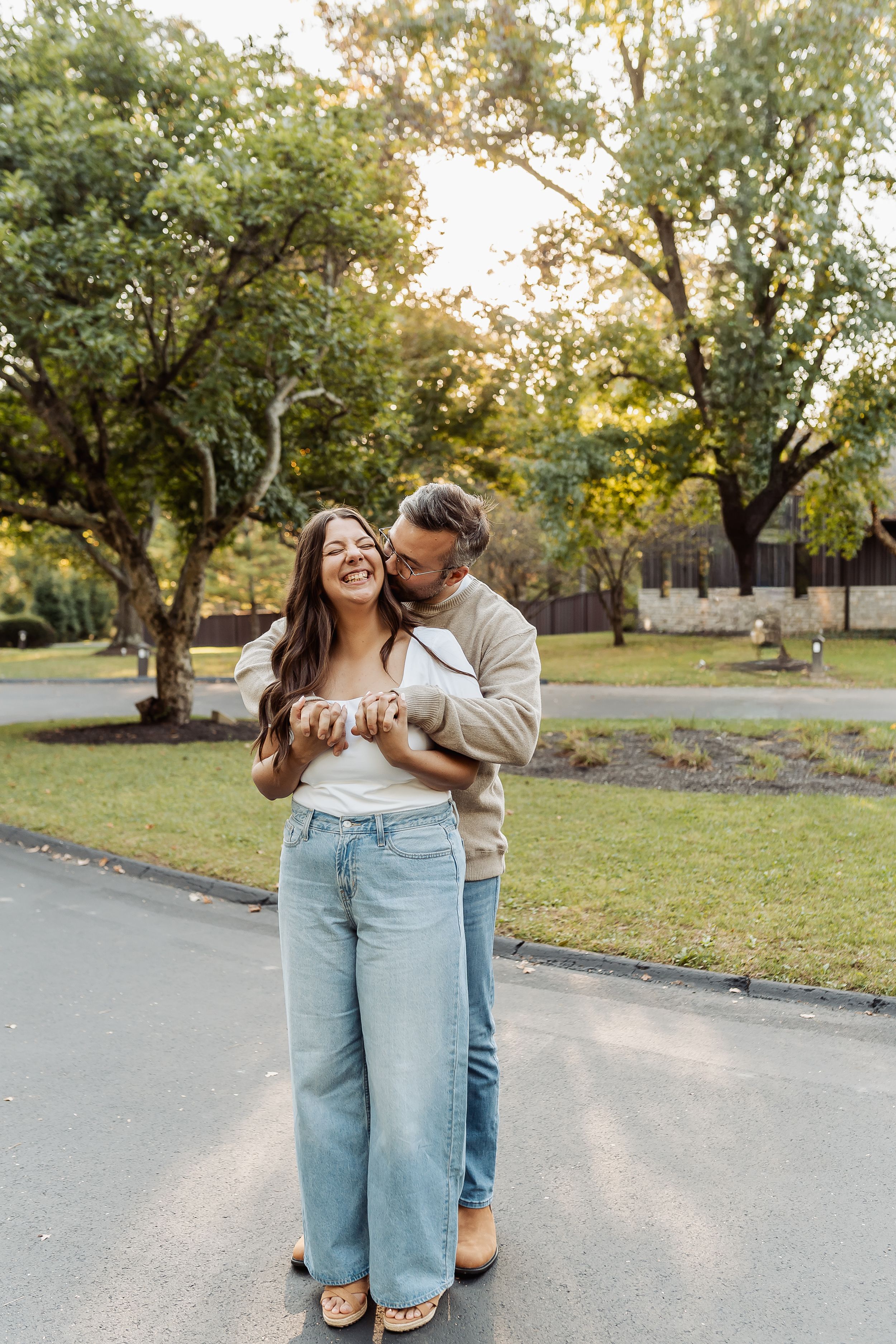 Happy couple embracing outdoors in a park, woman in wide-leg jeans laughing as man hugs her from behind.