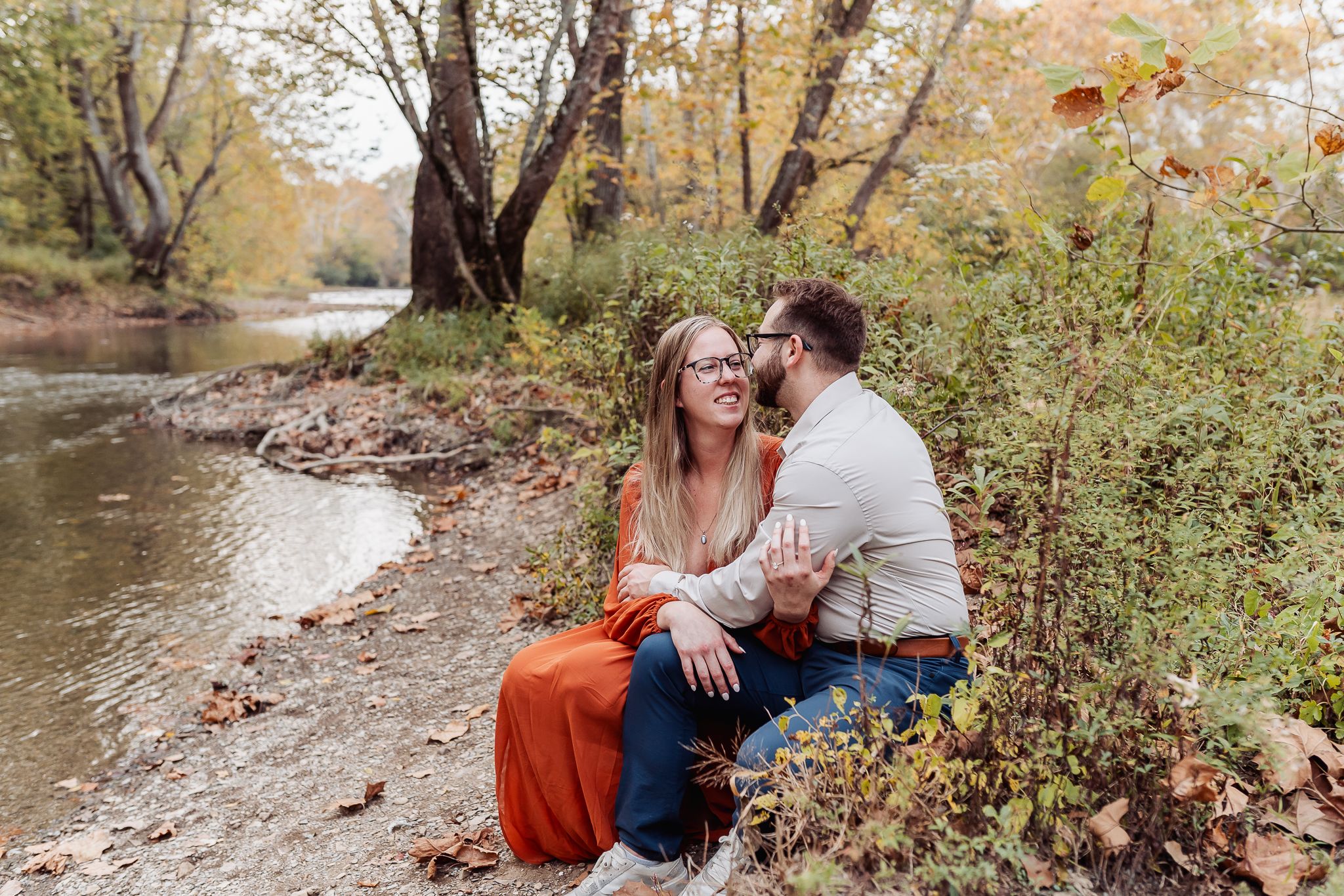 Couple sitting by a creek in autumn, woman in orange dress smiling as man whispers in her ear.
