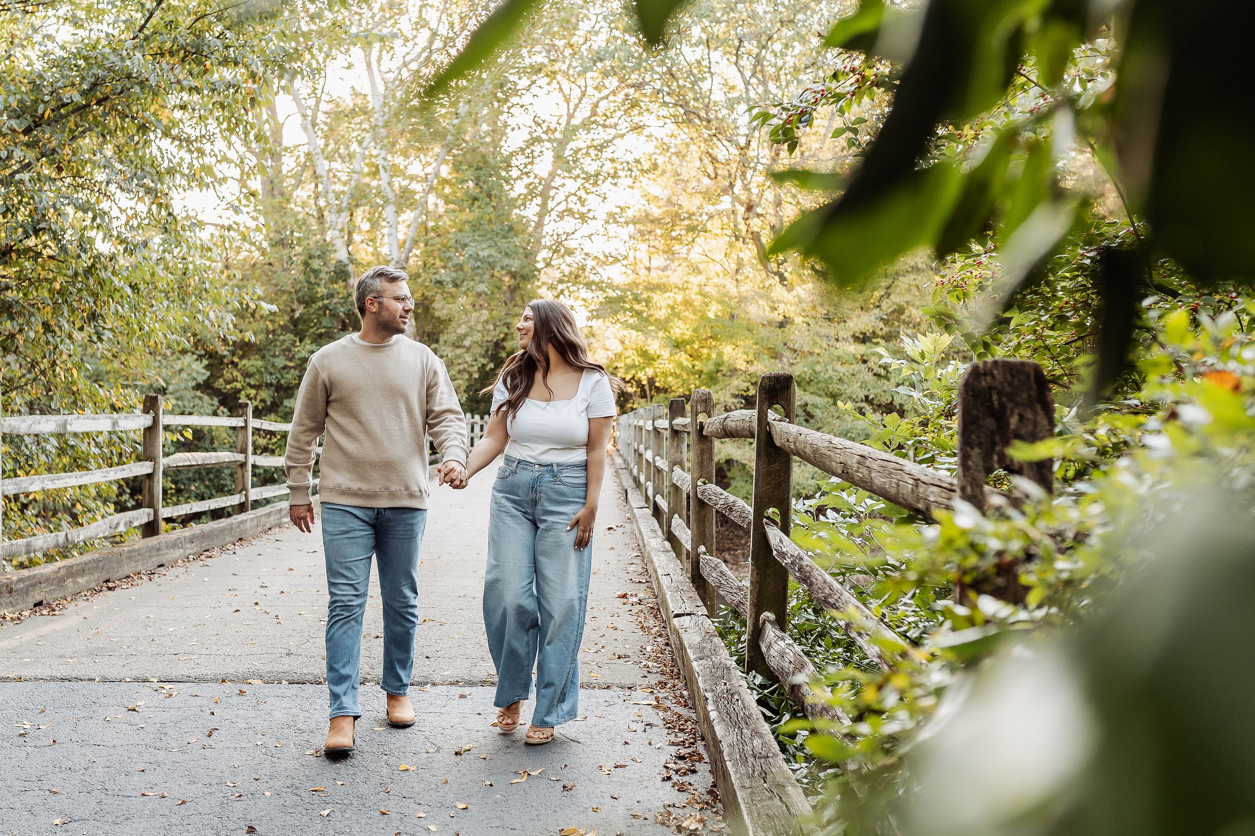 A couple walks hand-in-hand on a bridge surrounded by lush green trees in a park setting.