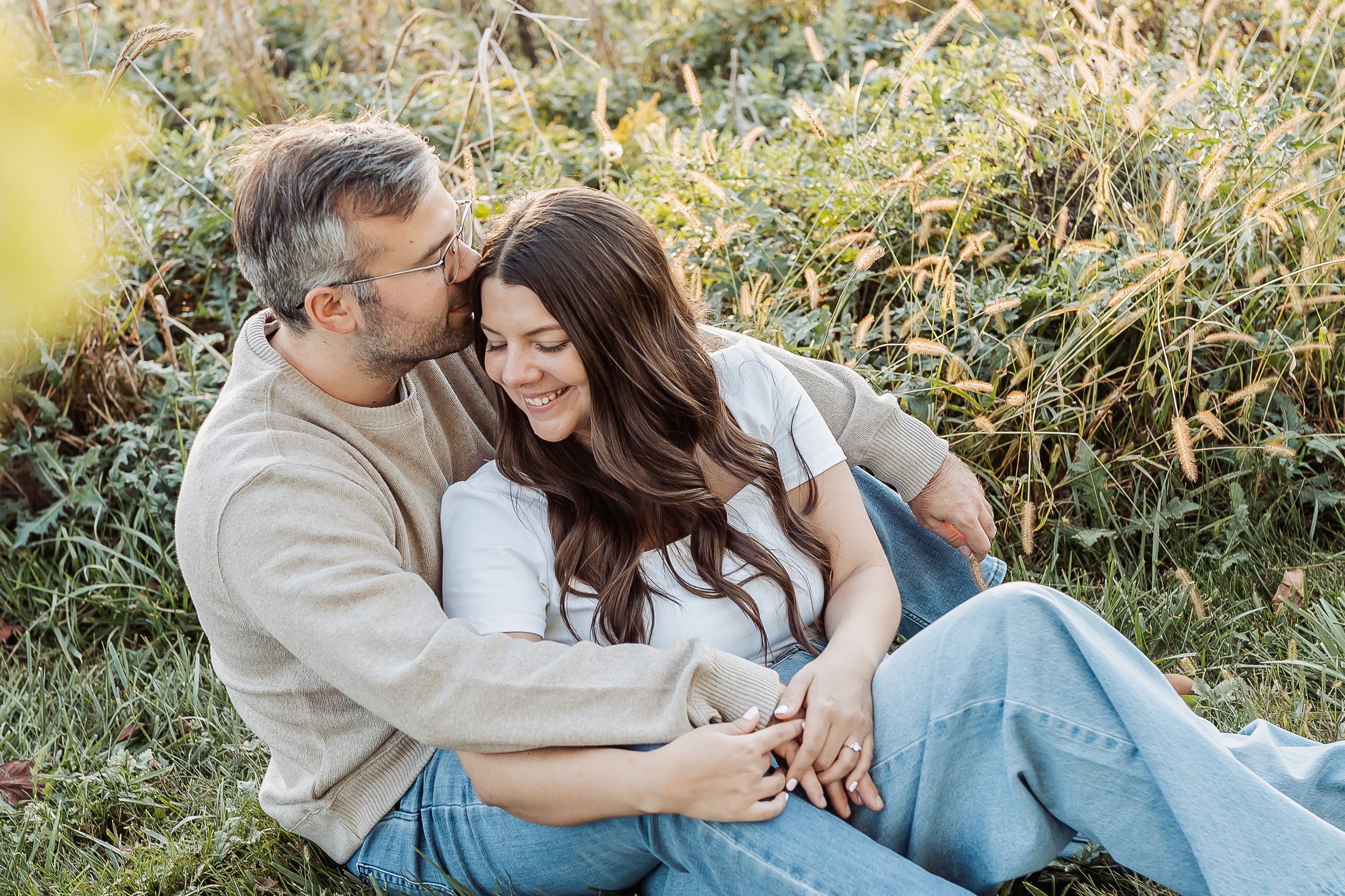 Couple sitting together in a field, smiling and embracing romantically during an outdoor engagement photo session.