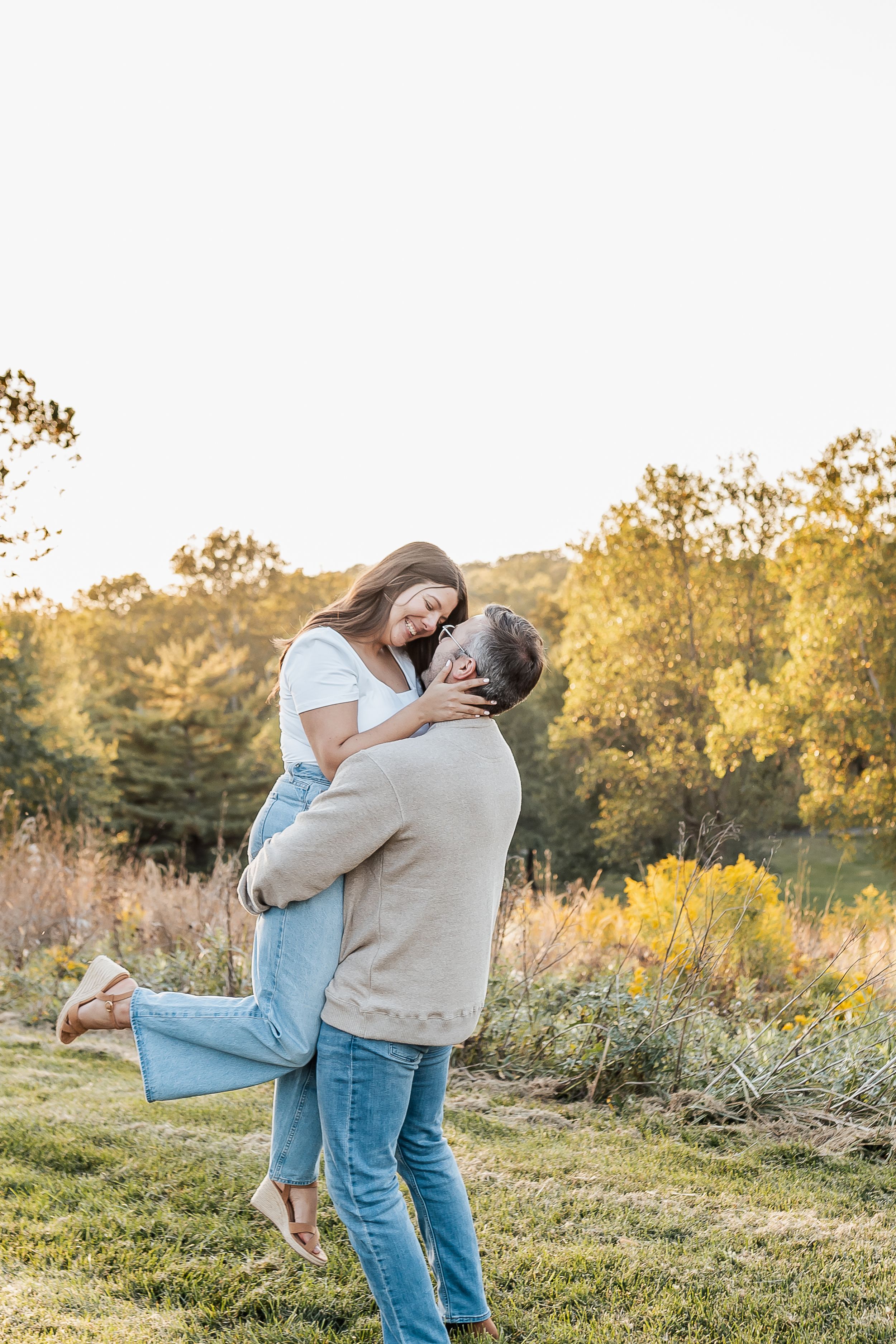 Couple embracing outdoors at golden hour, man lifting woman in jeans and white top, autumn trees in background.