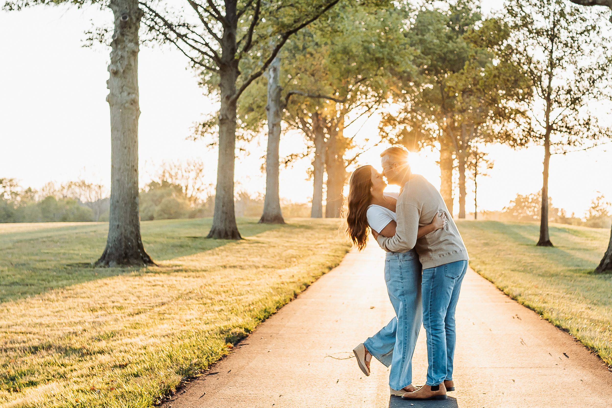 Couple sharing a romantic kiss on a sunlit park path surrounded by tall trees during golden hour sunset.