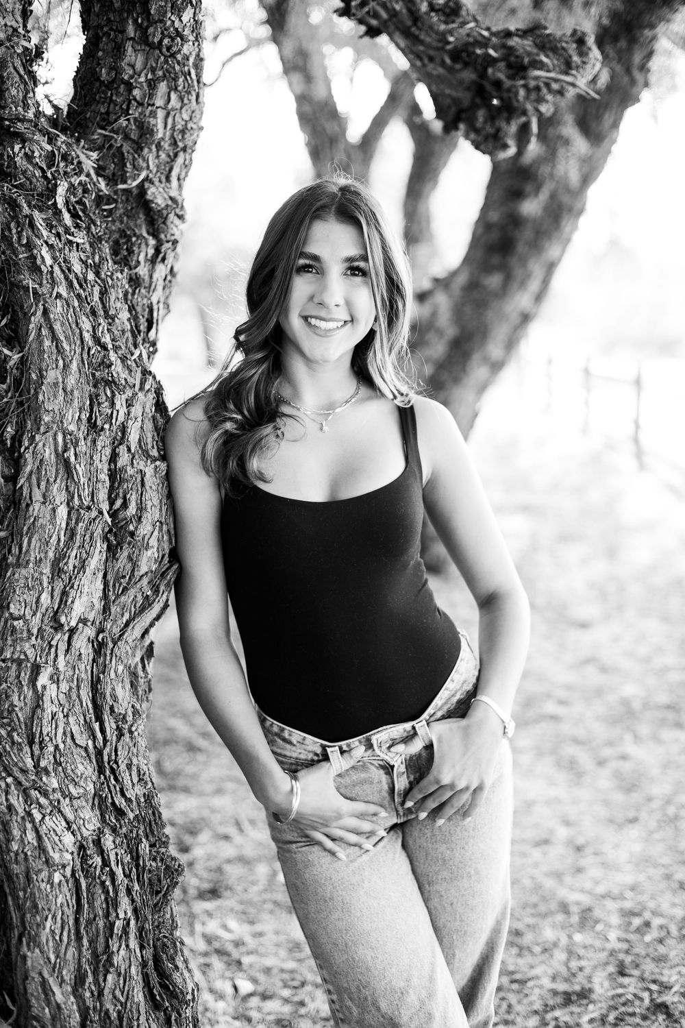 Black and white photo of a young woman in jeans and tank top leaning against a tree, smiling outdoors.