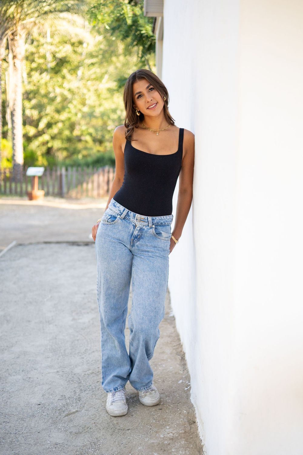 Young woman in black tank top and wide-leg jeans leaning against white wall outdoors, smiling casually.