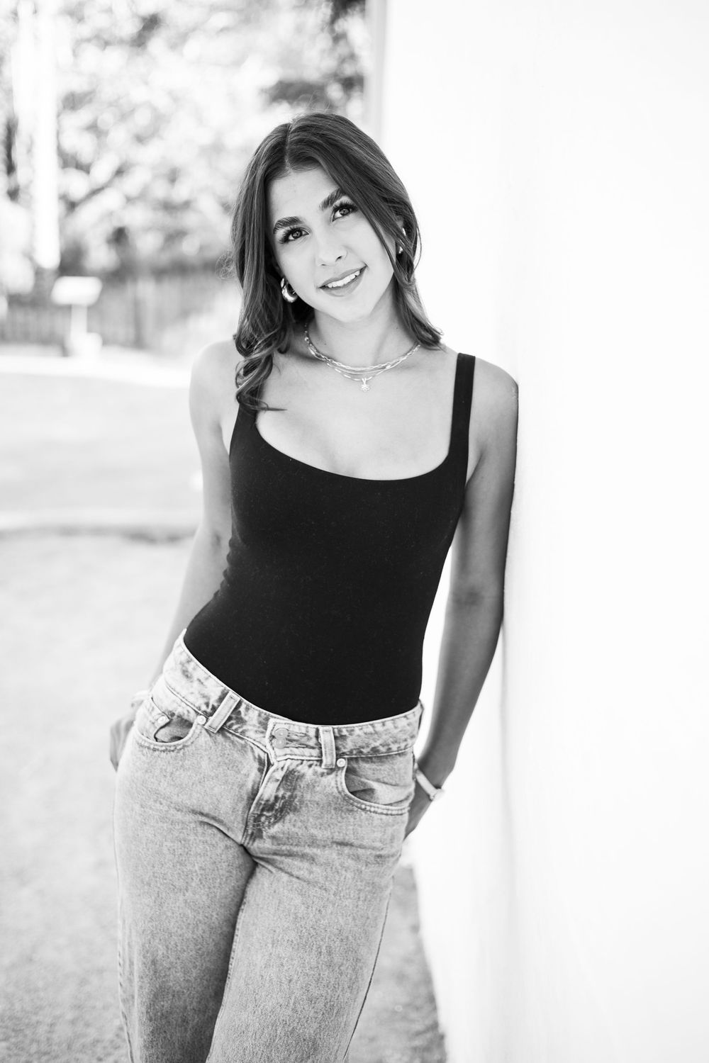 Black and white photo of a young woman in a black tank top and jeans, smiling and leaning against a white wall.