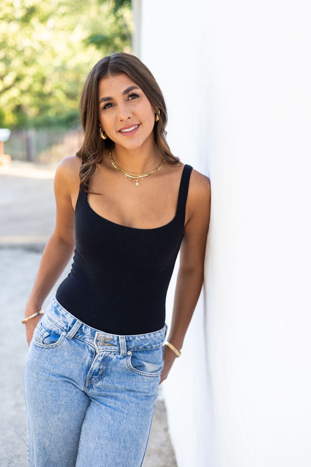 Young woman in black tank top and jeans leaning against white wall, wearing gold necklaces, smiling outdoors.