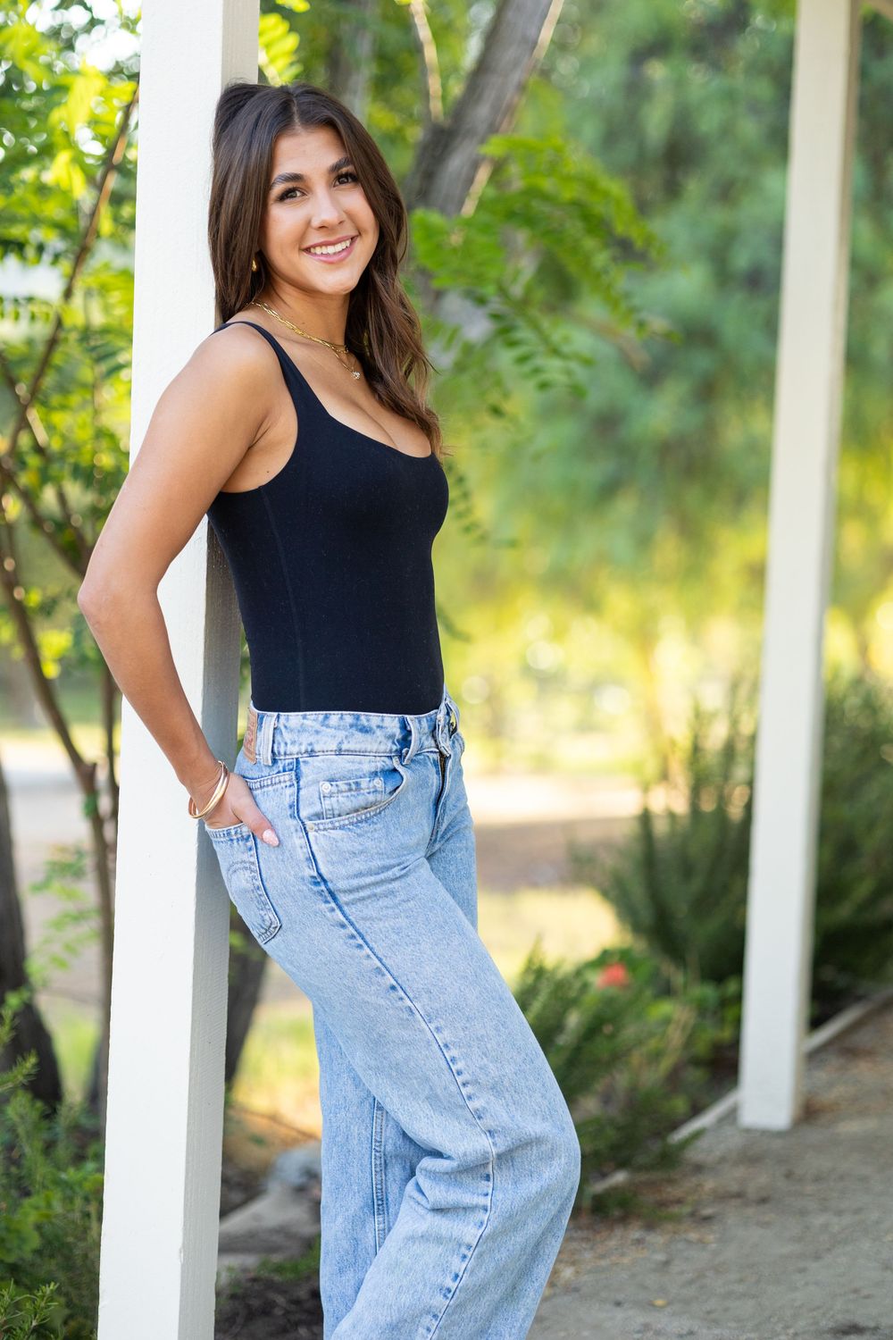 Young woman in black tank top and wide-leg jeans smiling outdoors, leaning against a white post surrounded by greenery.