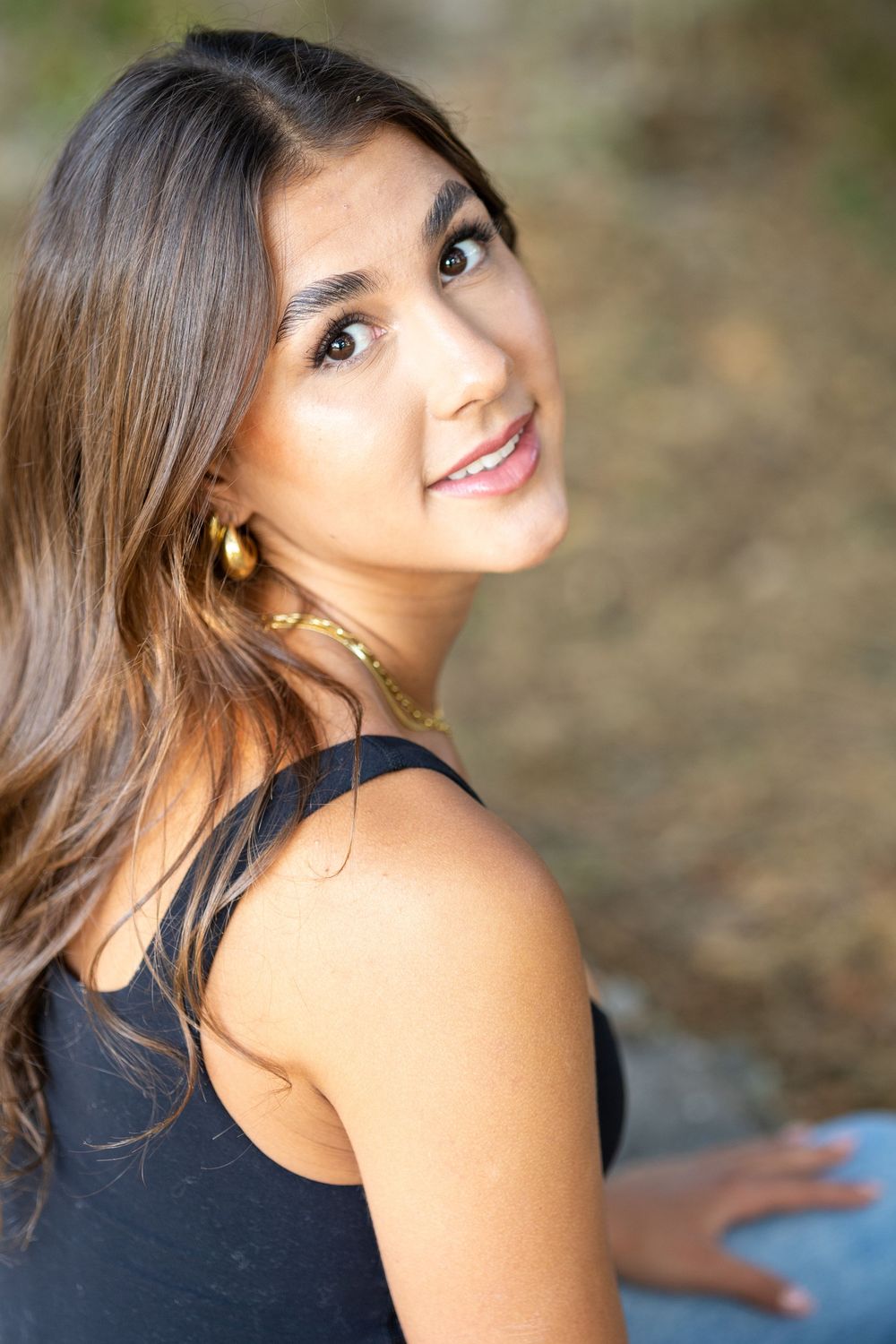 Young woman with long brown hair wearing gold jewelry and black top, smiling over shoulder outdoors.
