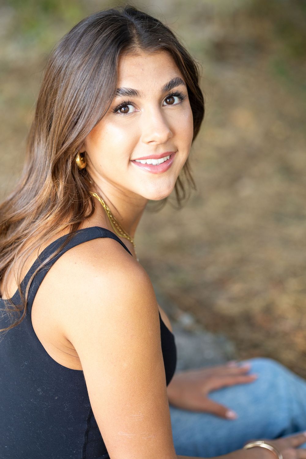 Young woman with long brown hair smiling over shoulder, wearing black top and gold necklace outdoors.