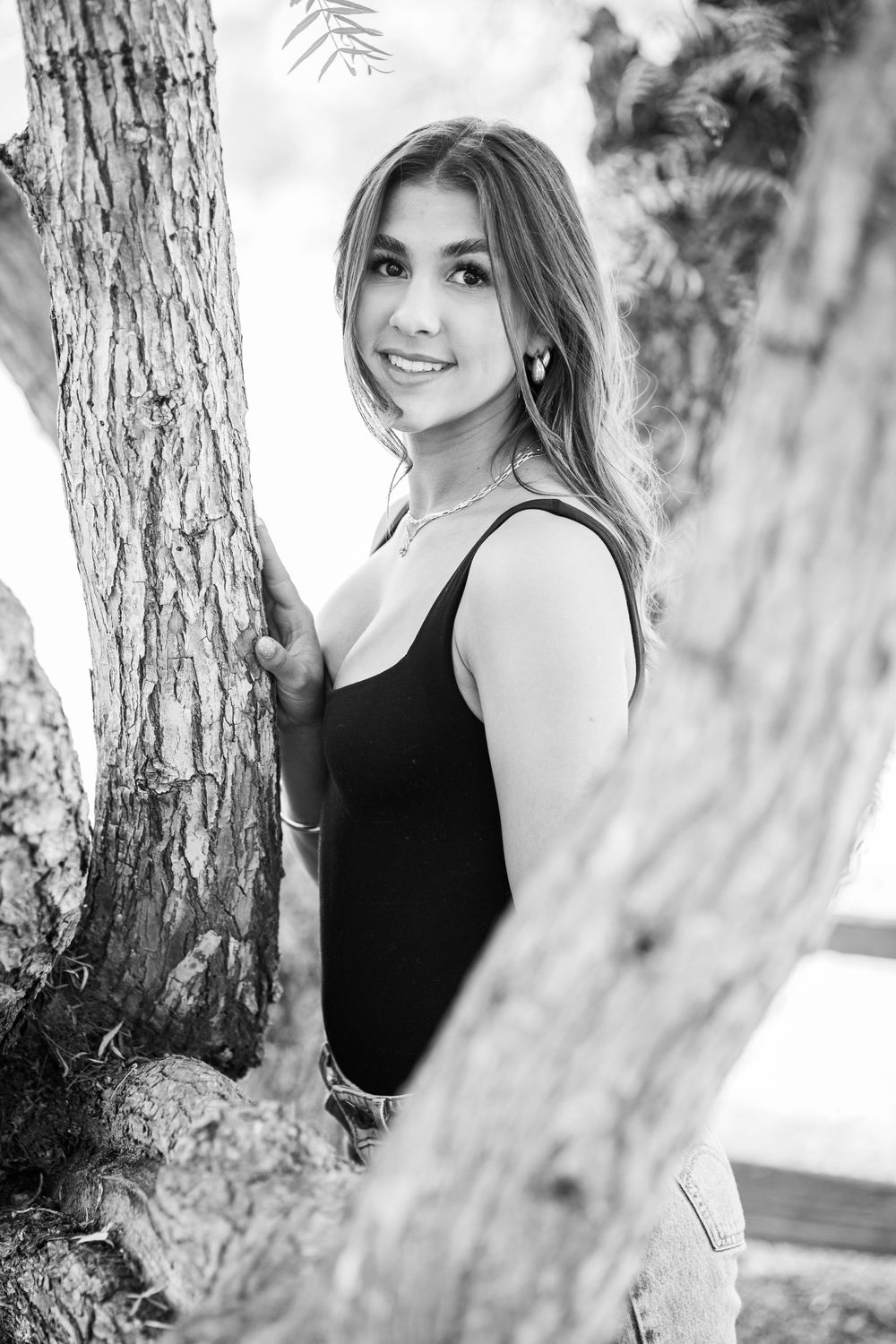 Black and white portrait of smiling young woman leaning against tree trunk in outdoor setting.