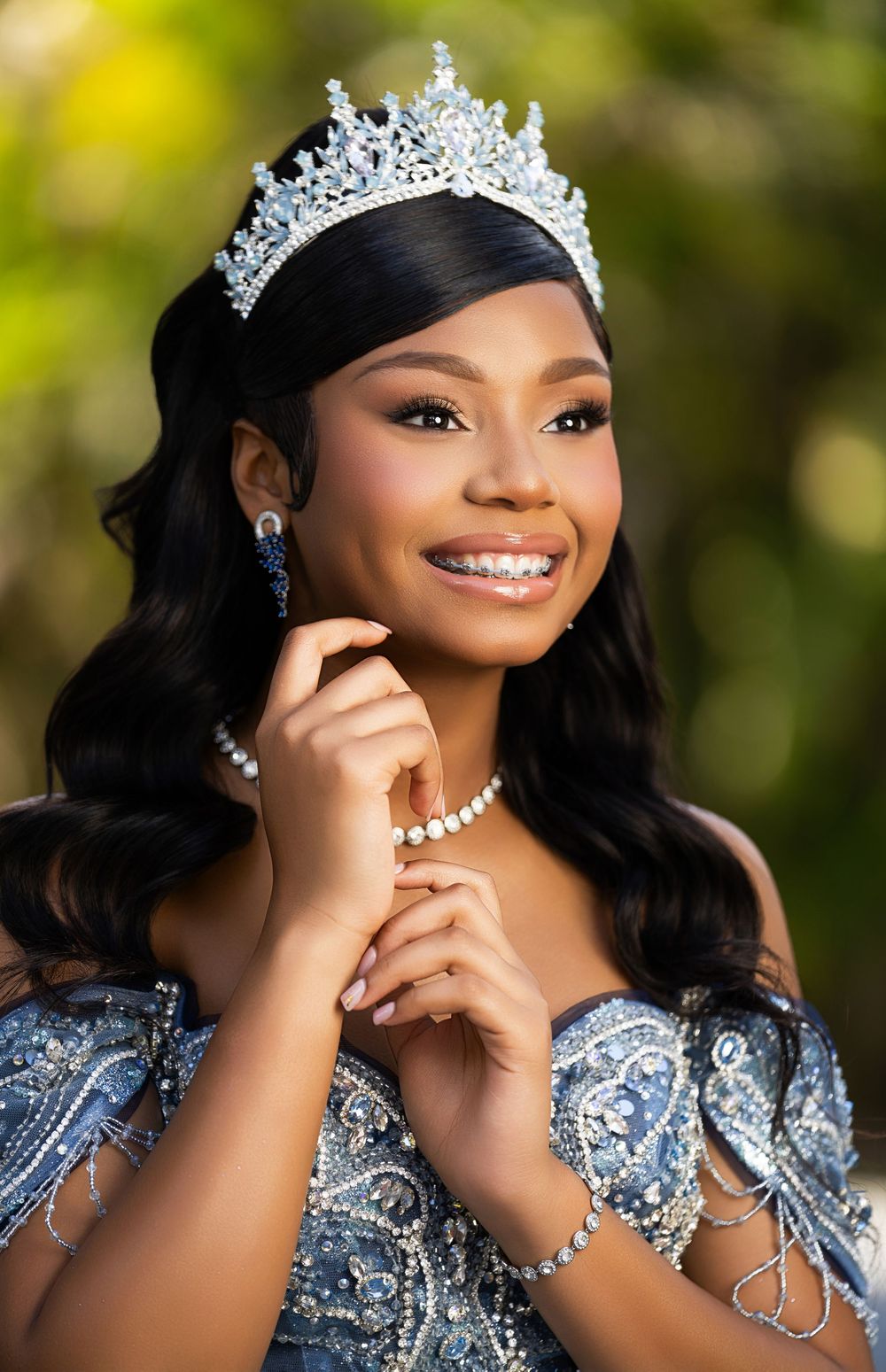 Smiling young woman wearing a sparkling tiara, blue beaded gown, and diamond jewelry, posing outdoors.