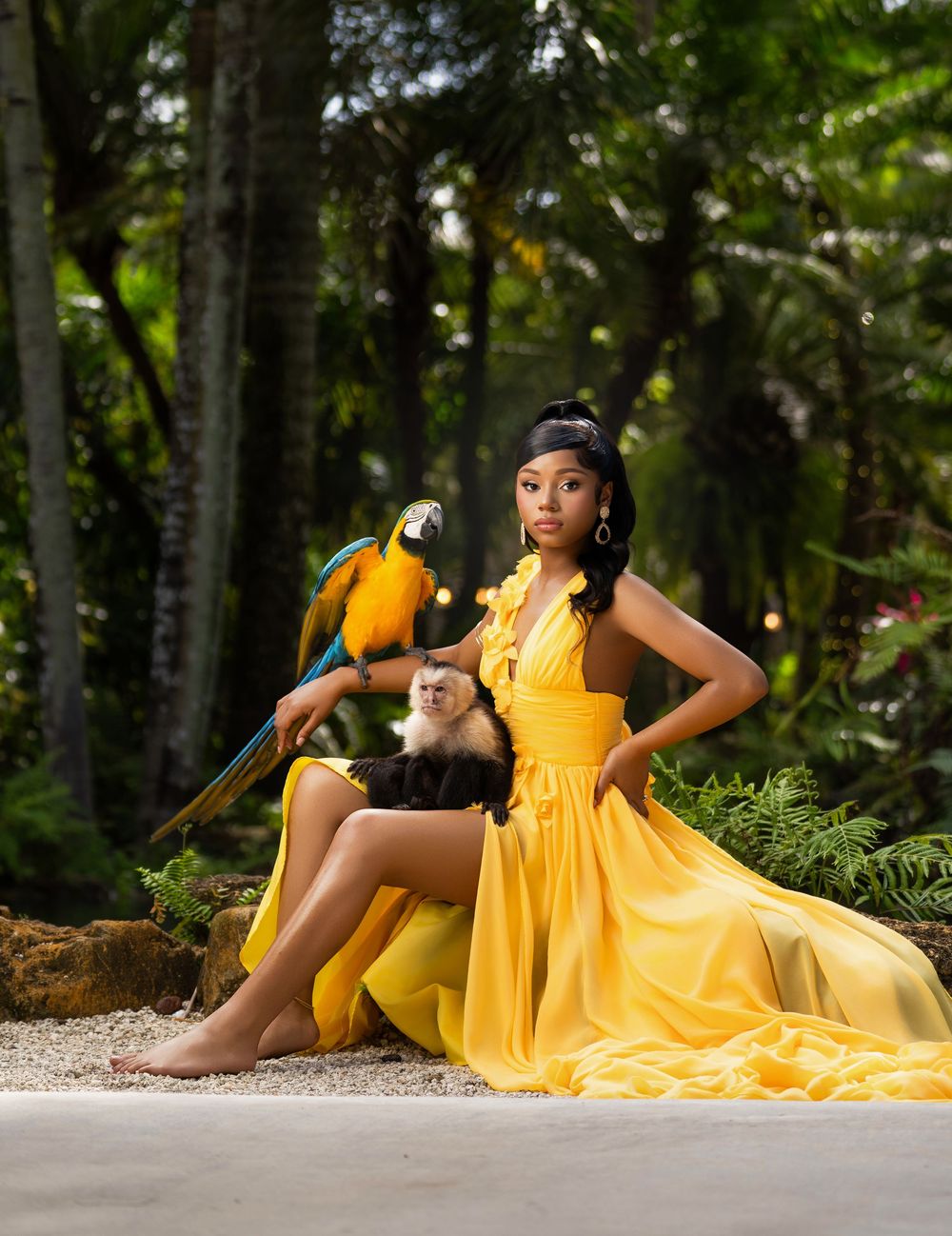 Woman in yellow gown poses with blue-gold macaw and white capuchin monkey in lush tropical forest setting.