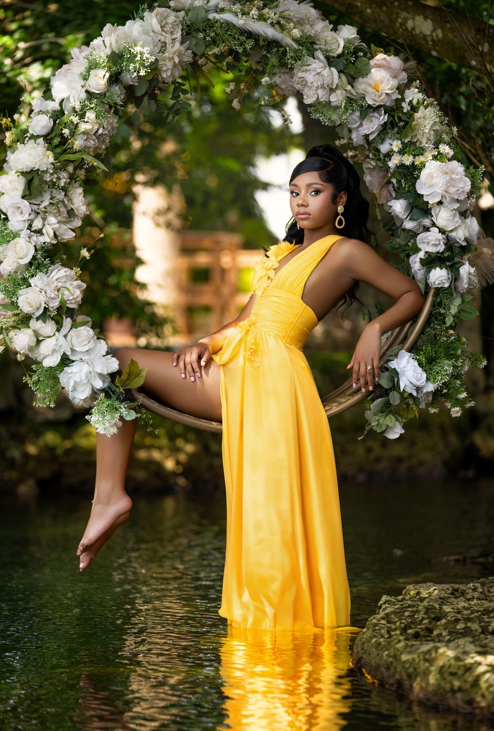 A woman in a yellow gown poses on a floral swing arch over water in a lush garden setting.