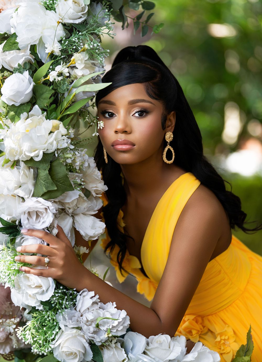 Young woman in yellow dress poses elegantly among white roses, wearing gold earrings and a ring in a lush garden setting.