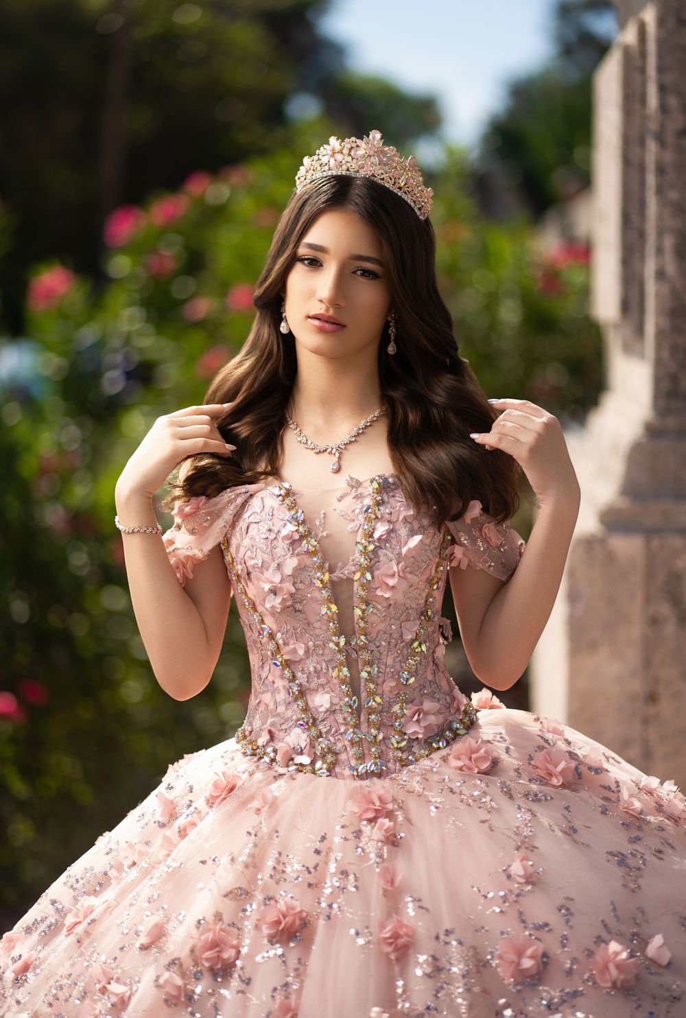 Young woman in pink floral ball gown with tiara and jewels poses outdoors for quinceañera portrait.