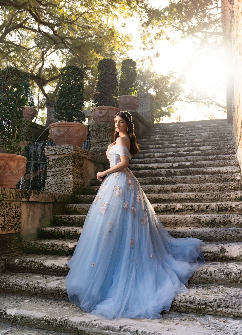 Woman in blue floral ball gown on stone steps in sunlit garden.
