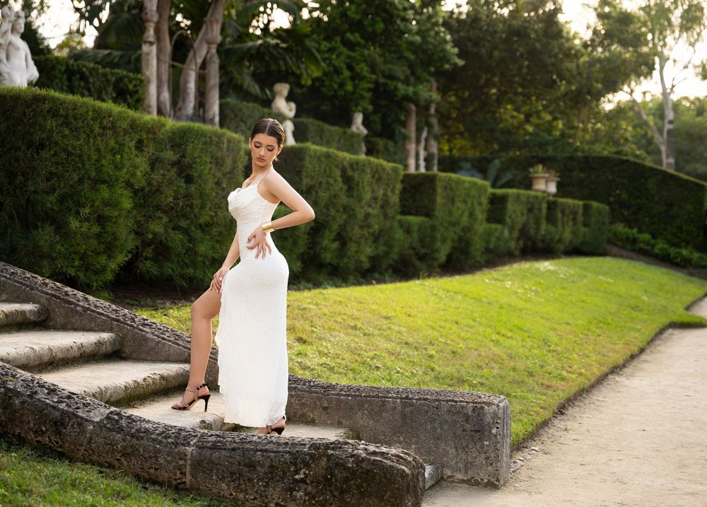 Woman in white gown poses on stone steps in formal garden with trimmed hedges.