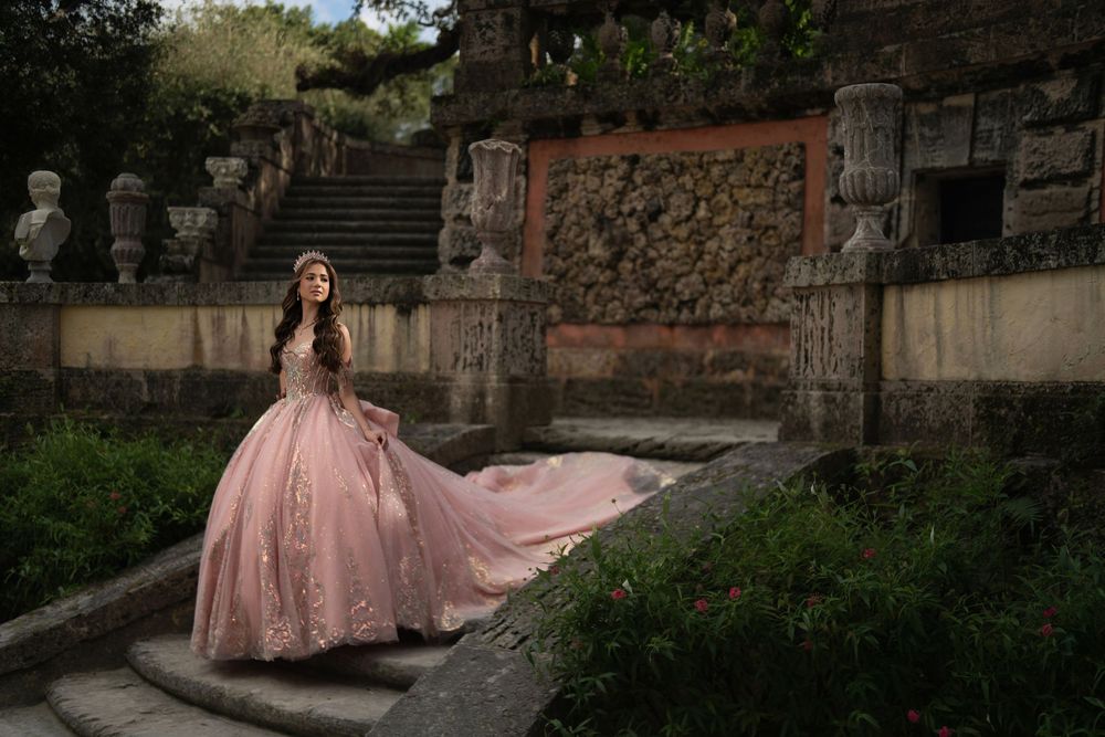 Young woman in pink ball gown poses gracefully in a stone garden setting for a quinceañera portrait.