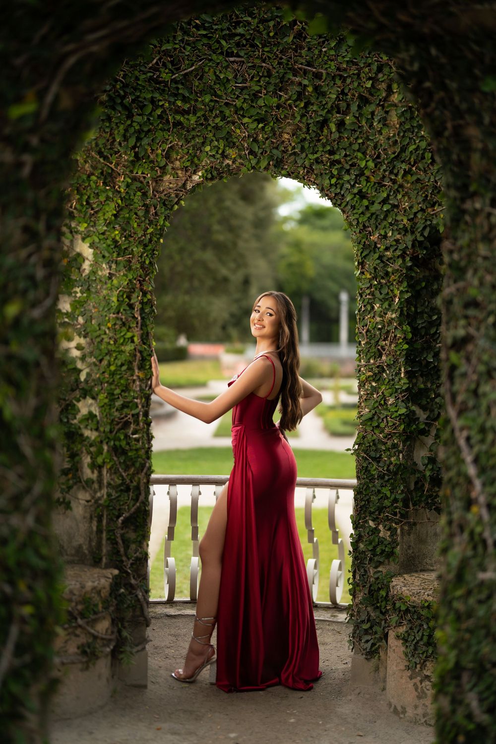 Young woman in elegant red gown poses gracefully under an ivy-covered stone arch in a lush garden setting.