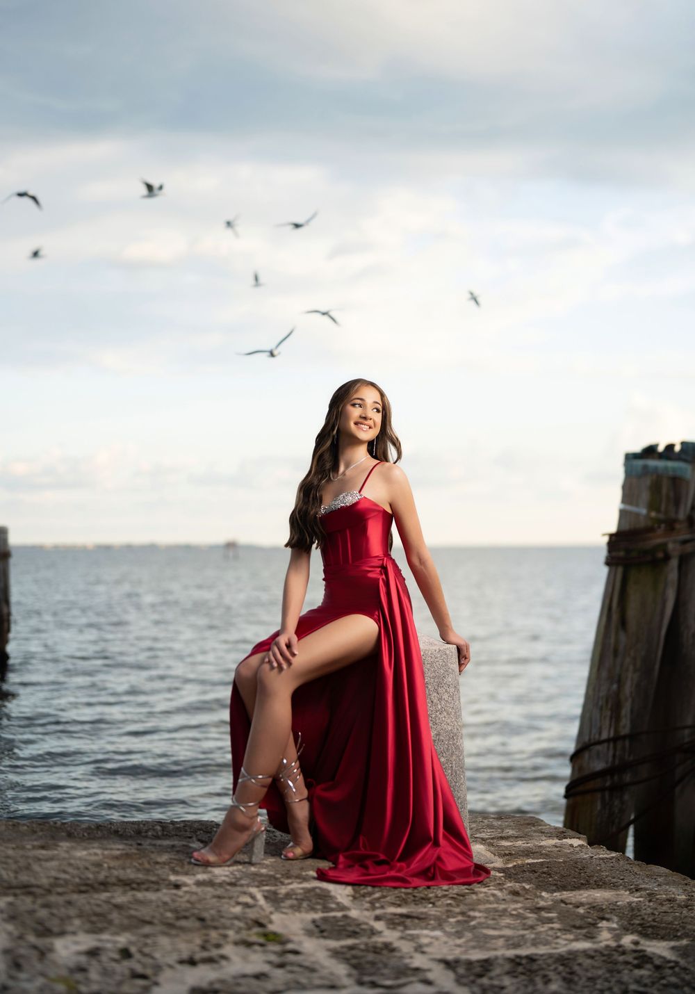 Young woman in red satin gown poses on seaside pier with seagulls flying overhead against cloudy sky.