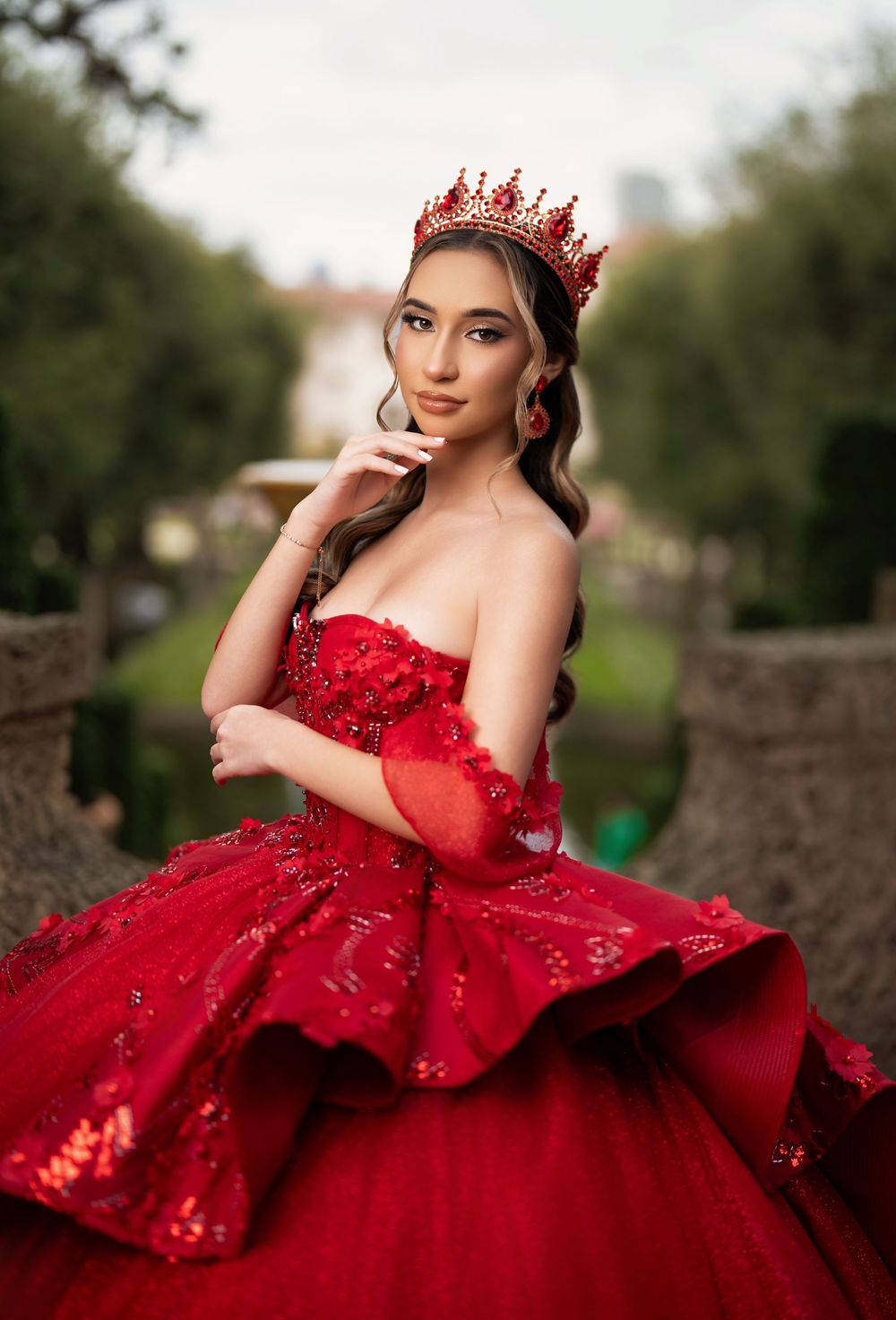 Young woman in red ballgown and crown poses elegantly outdoors for quinceañera portrait.