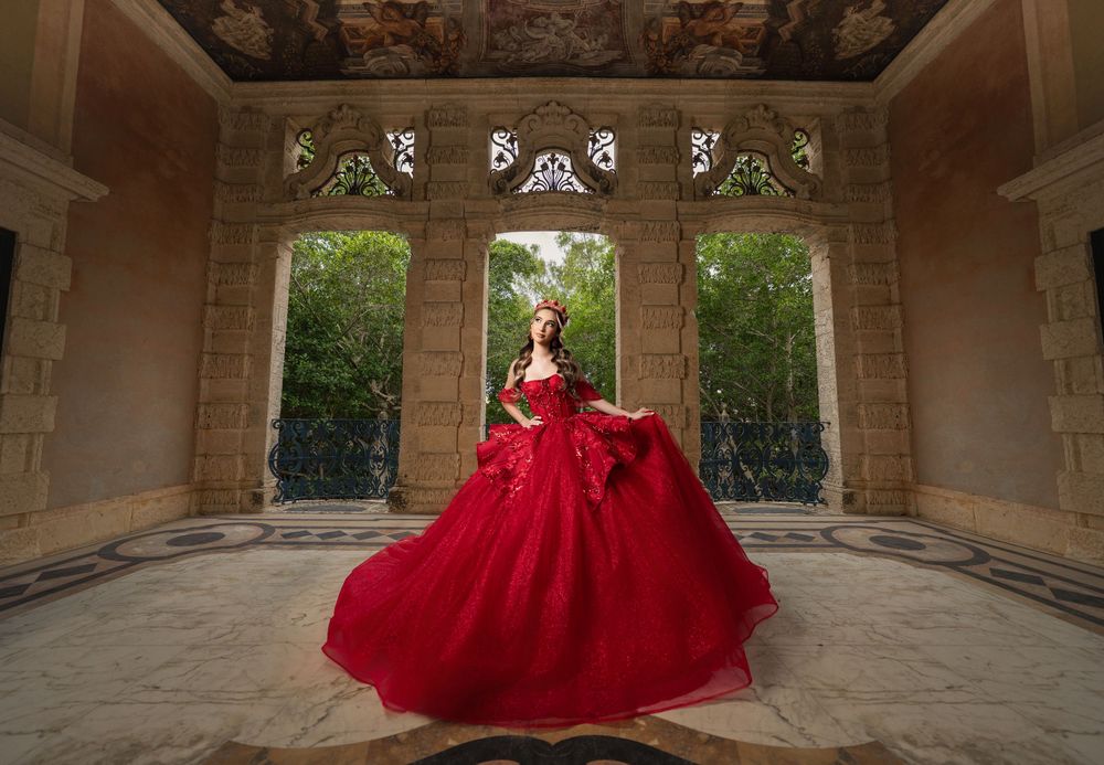 Woman in voluminous red ball gown poses in elegant stone pavilion with arched windows and lush garden backdrop.