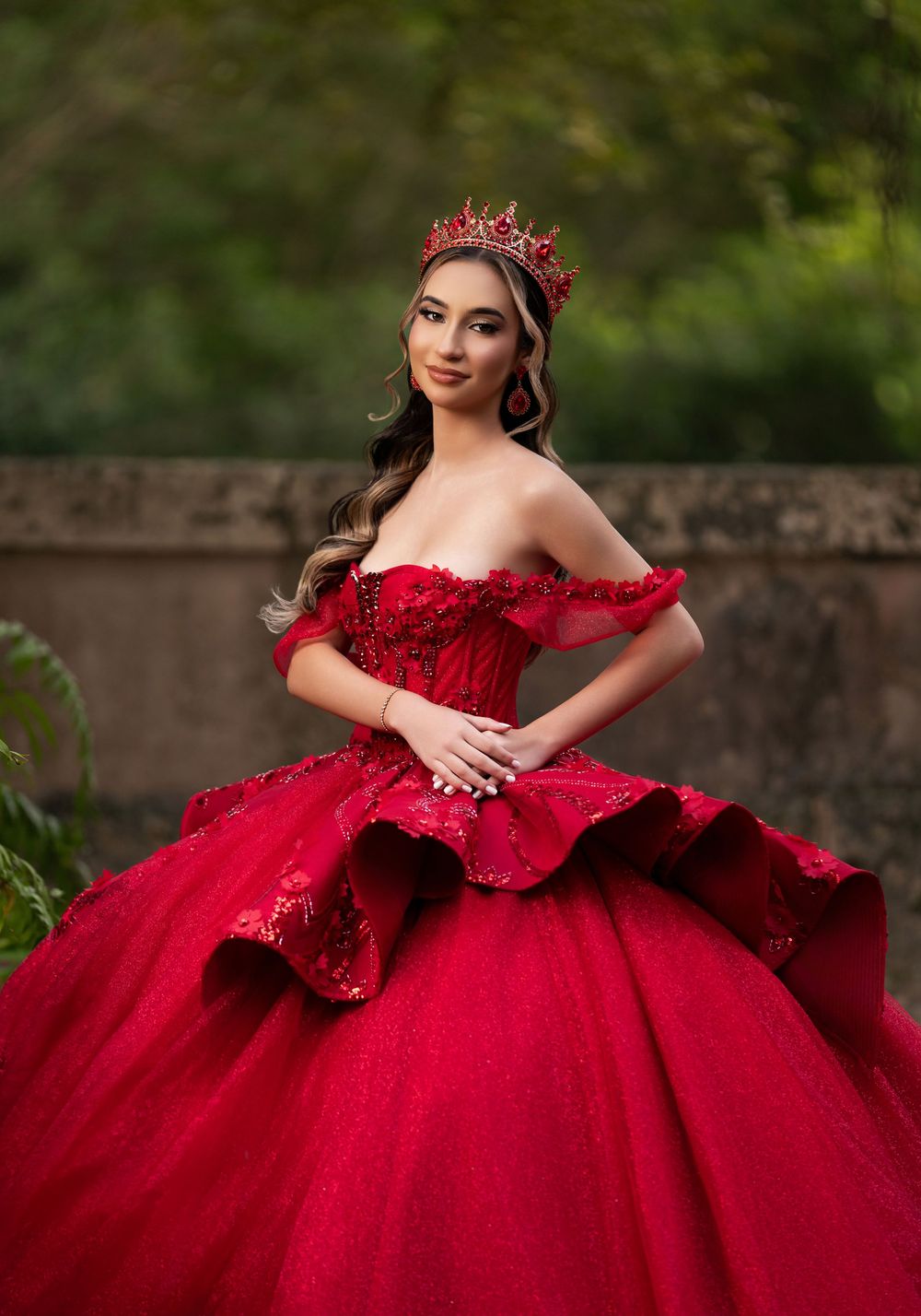 Young woman in red off-shoulder ball gown with crown poses outdoors for quinceañera portrait.