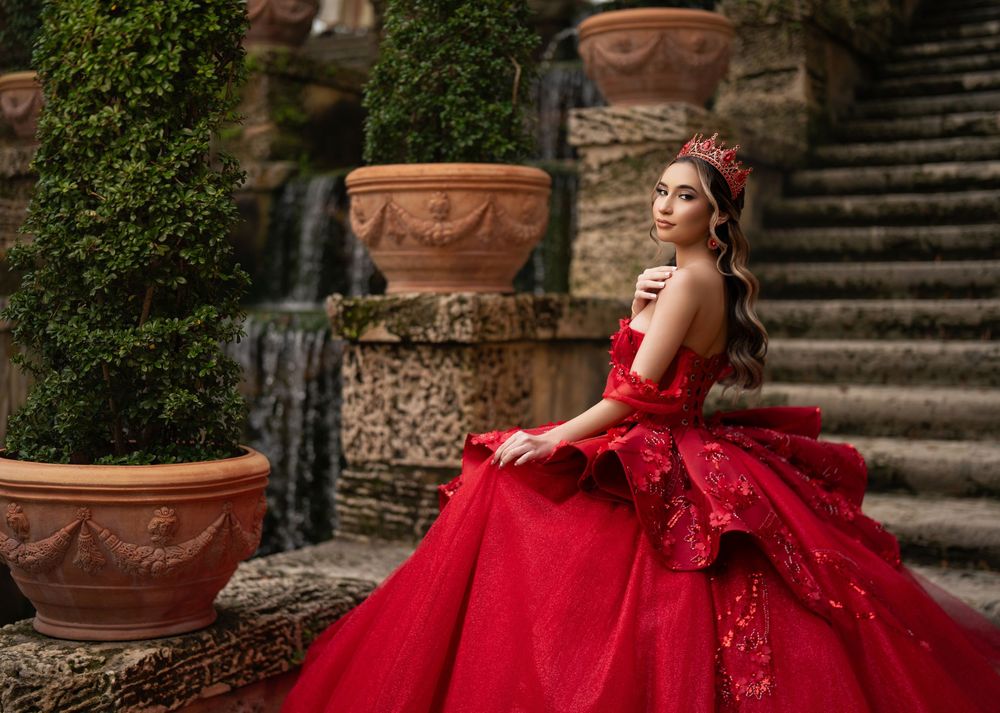 Young woman in a stunning red ball gown and crown poses gracefully on stone steps surrounded by lush greenery and terracotta pots.