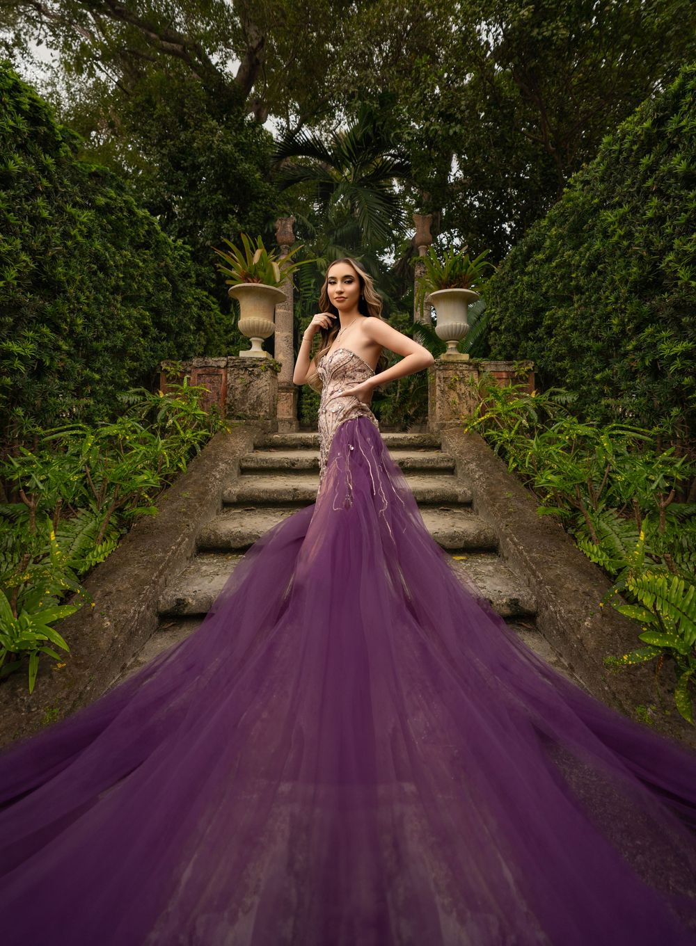 Model in purple tulle mermaid gown with long train stands on garden stairway.