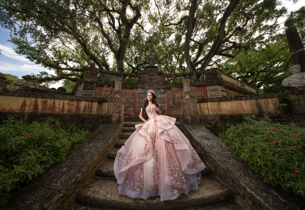 Young woman in a stunning pink ball gown poses on stone steps surrounded by lush greenery for quinceañera photos.