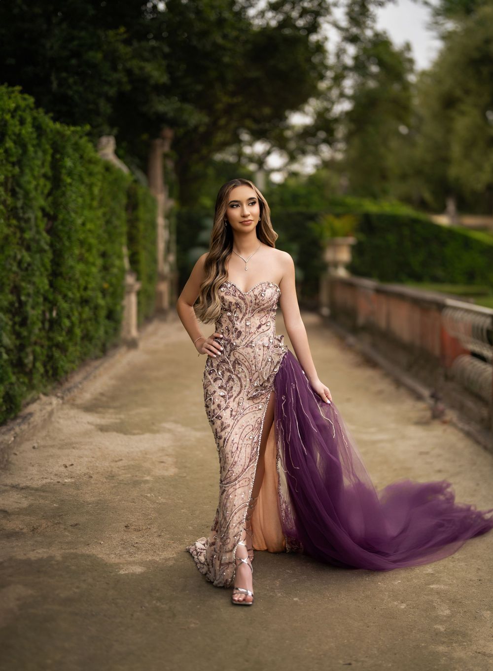 Young woman in sequined mermaid gown with purple tulle train poses on garden pathway surrounded by greenery.