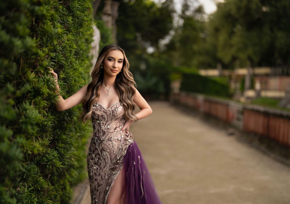 Young woman in elegant purple and gold sequin gown poses gracefully outdoors near lush green hedges.