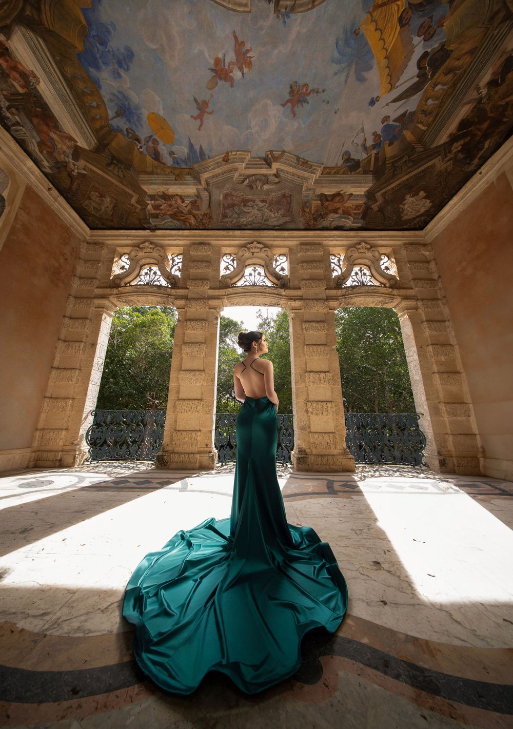 Woman in emerald green gown with train poses in ornate baroque pavilion with painted ceiling.