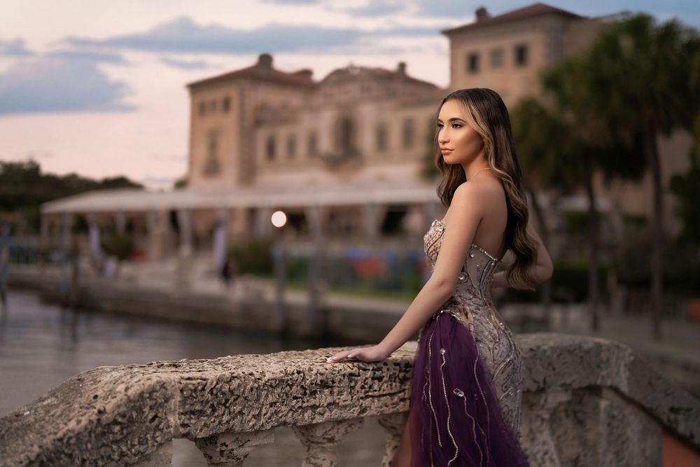 Elegant woman in purple beaded gown posing on stone bridge at dusk with historic building in background.