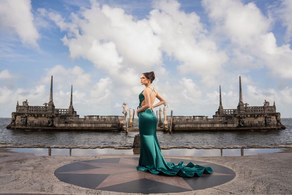Woman in elegant green mermaid gown poses at waterfront plaza with historic stone fortress in background.