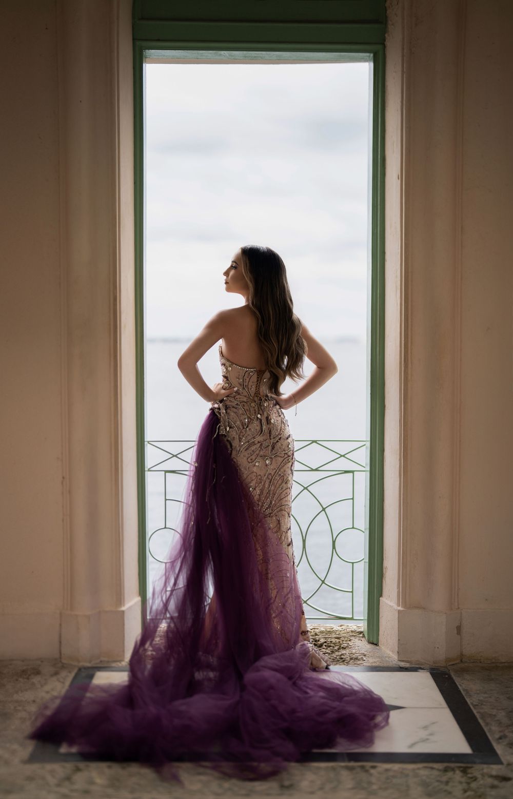 Woman in beaded purple tulle gown stands in doorway overlooking ocean view.