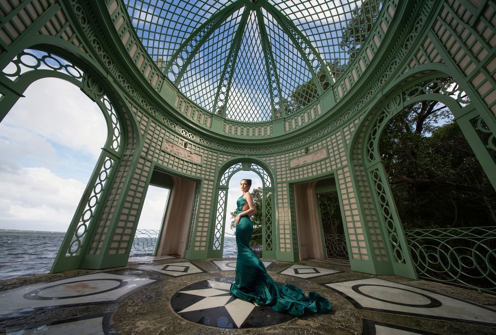 Woman in emerald green gown poses inside ornate green lattice pavilion overlooking water.