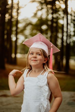 A young graduate in white dress and pink cap poses playfully in a forest setting.