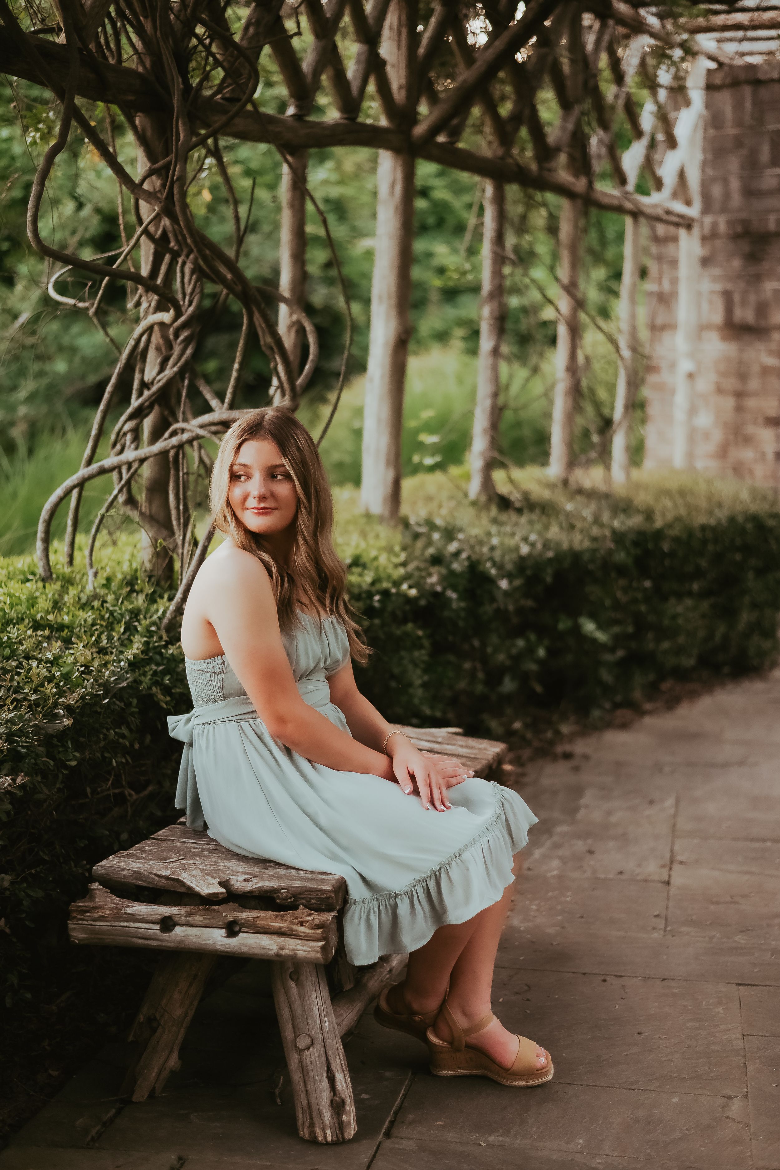 Person in mint green dress poses on rustic wooden bench under vine-covered pergola.