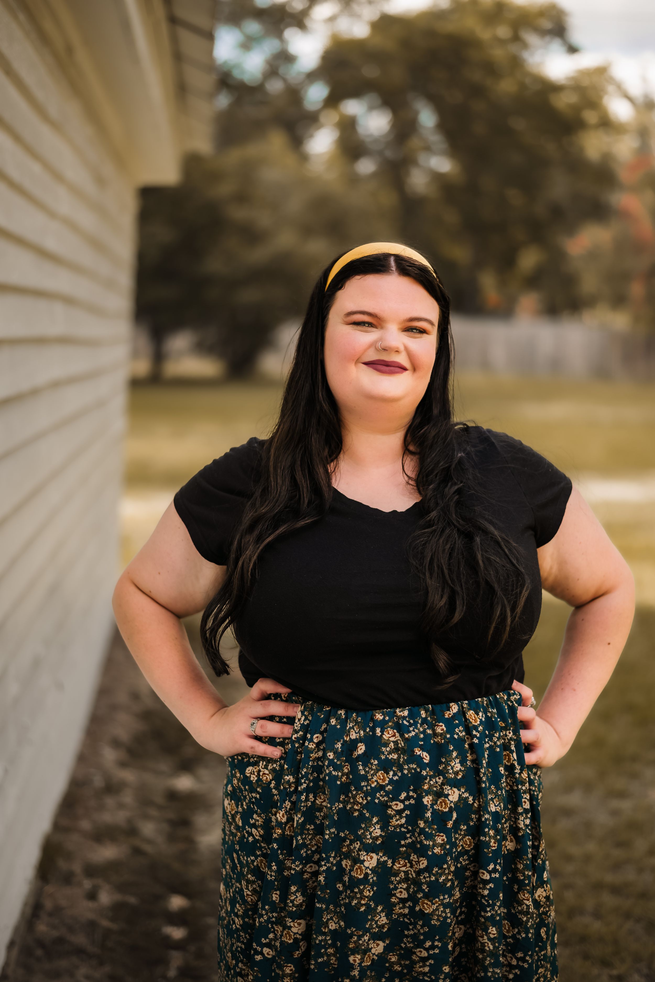 Casual outdoor portrait of a person in black top and floral skirt posing against a white wall.