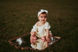 A baby in white dress and bow headband sitting on a mirror outdoors.