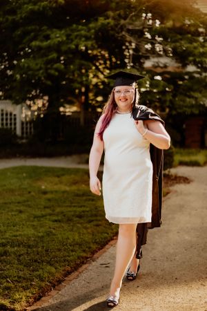 A person in a white dress walks along a sunlit path through a garden setting.