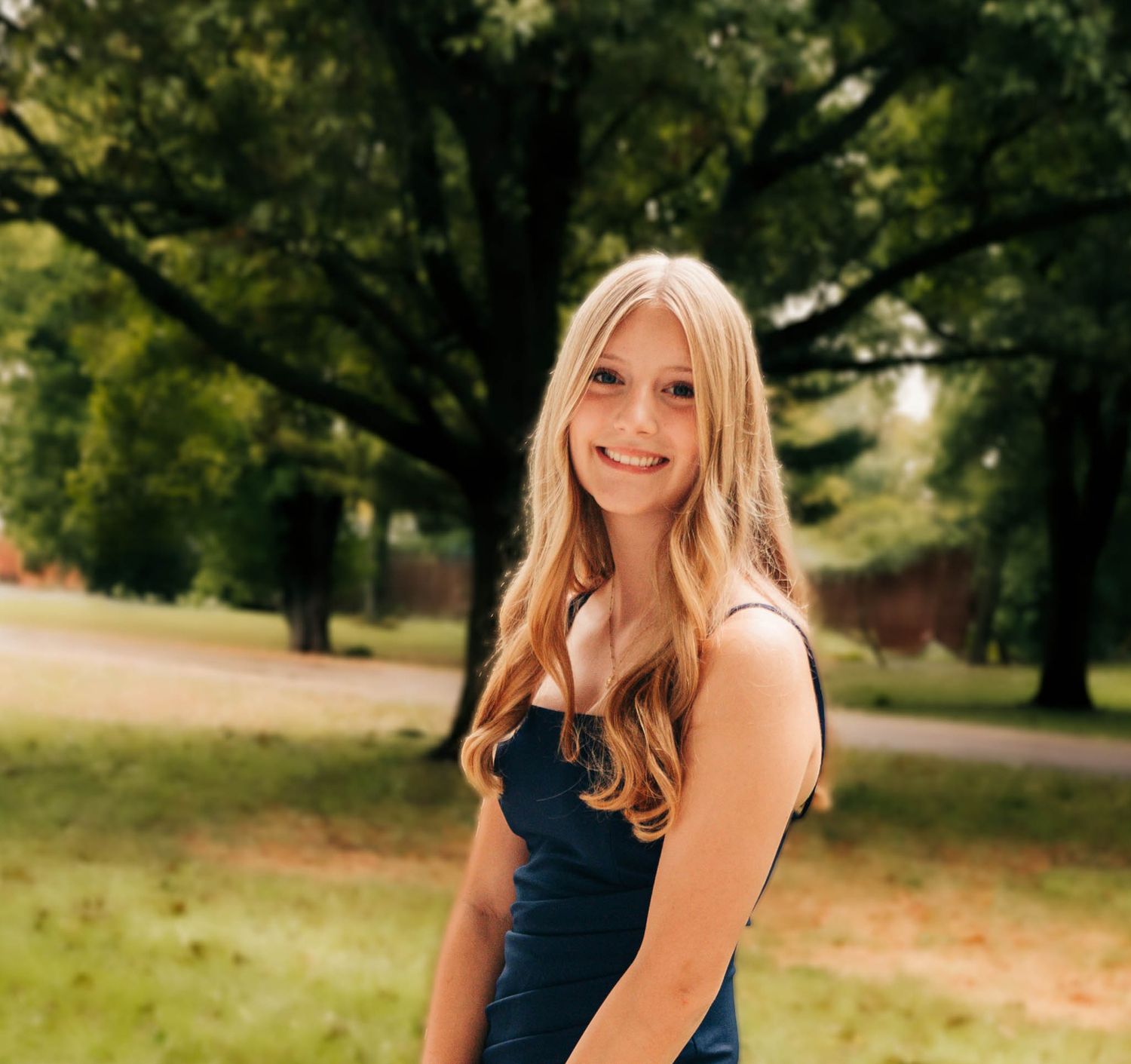 Woman in navy blue dress poses outdoors in park setting with natural greenery.