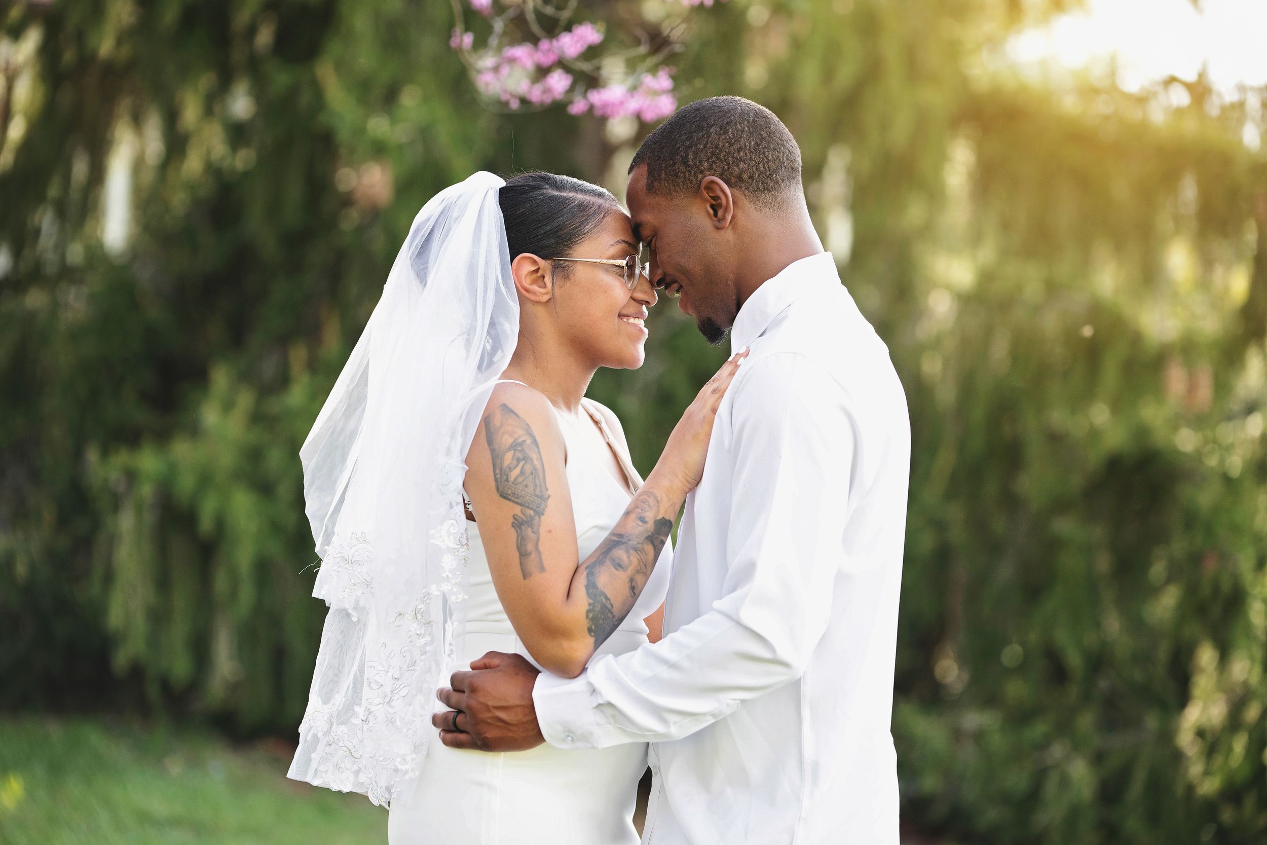 A couple in wedding attire embraces outdoors with lush greenery in the background.
