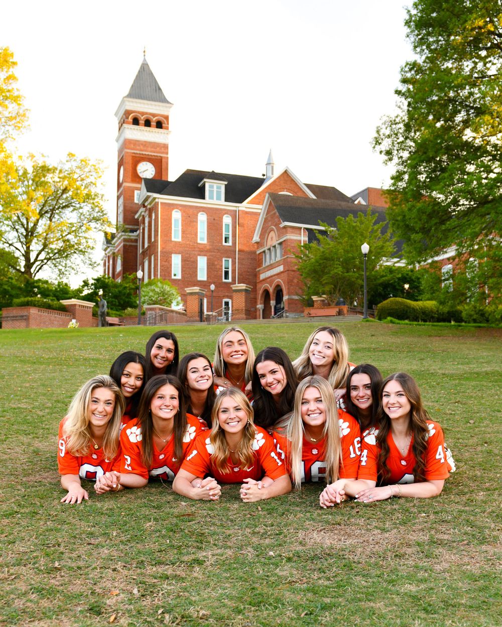 Group of people in matching orange shirts lying on grass in front of brick building with clock tower.