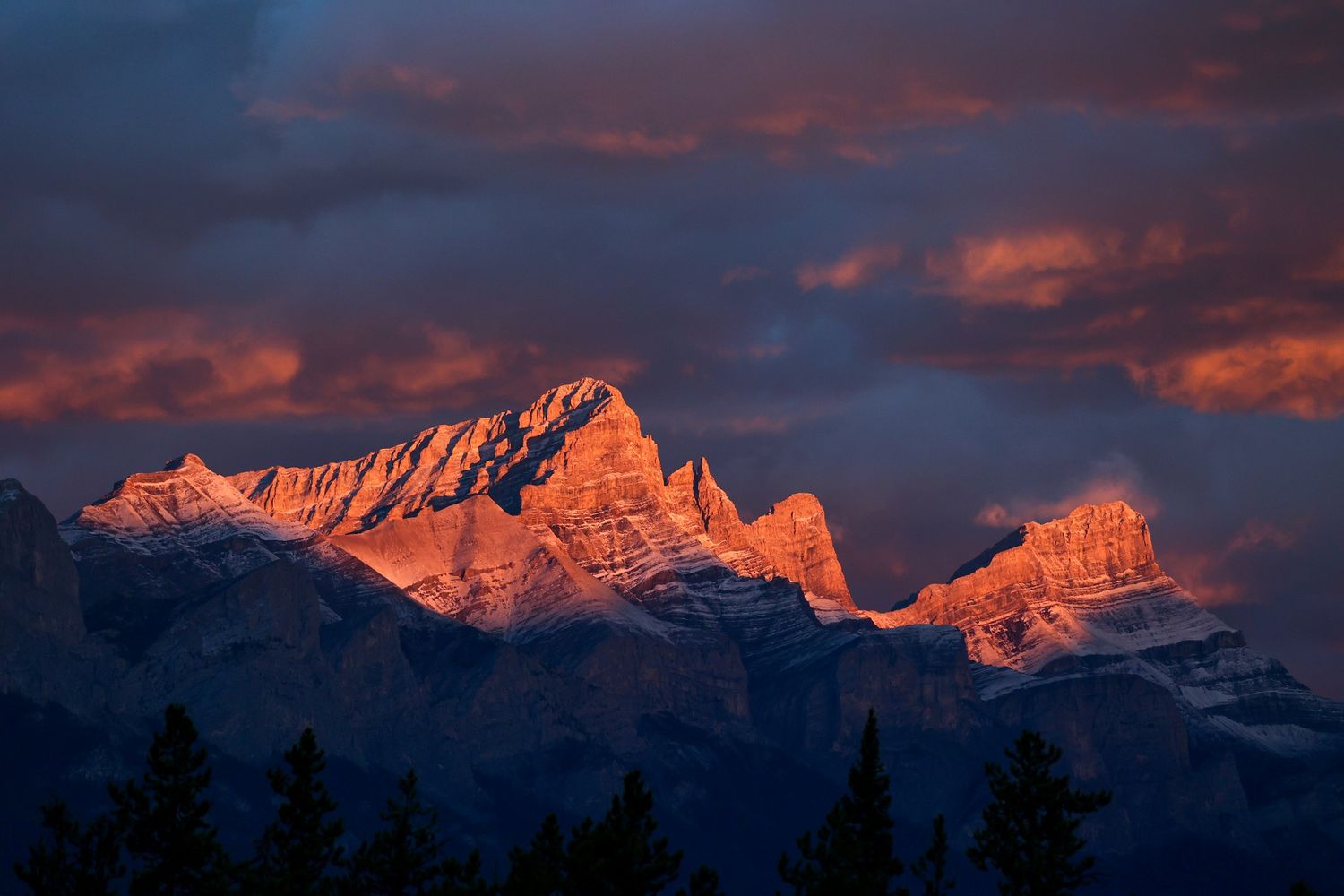Dramatic alpenglow illuminates snow-capped mountain peaks under a stormy purple and orange sunset sky.