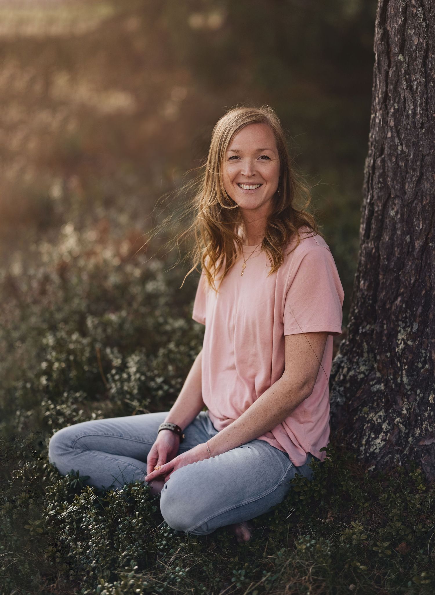 Smiling young blonde woman in pink shirt sitting cross-legged against a tree in golden forest light.