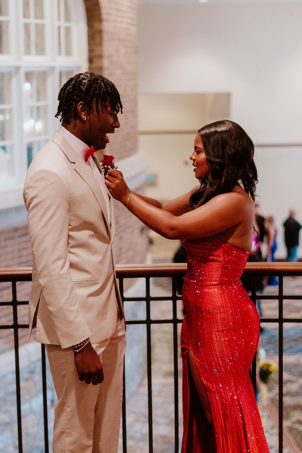 Person in red dress adjusting boutonniere on person wearing beige suit.