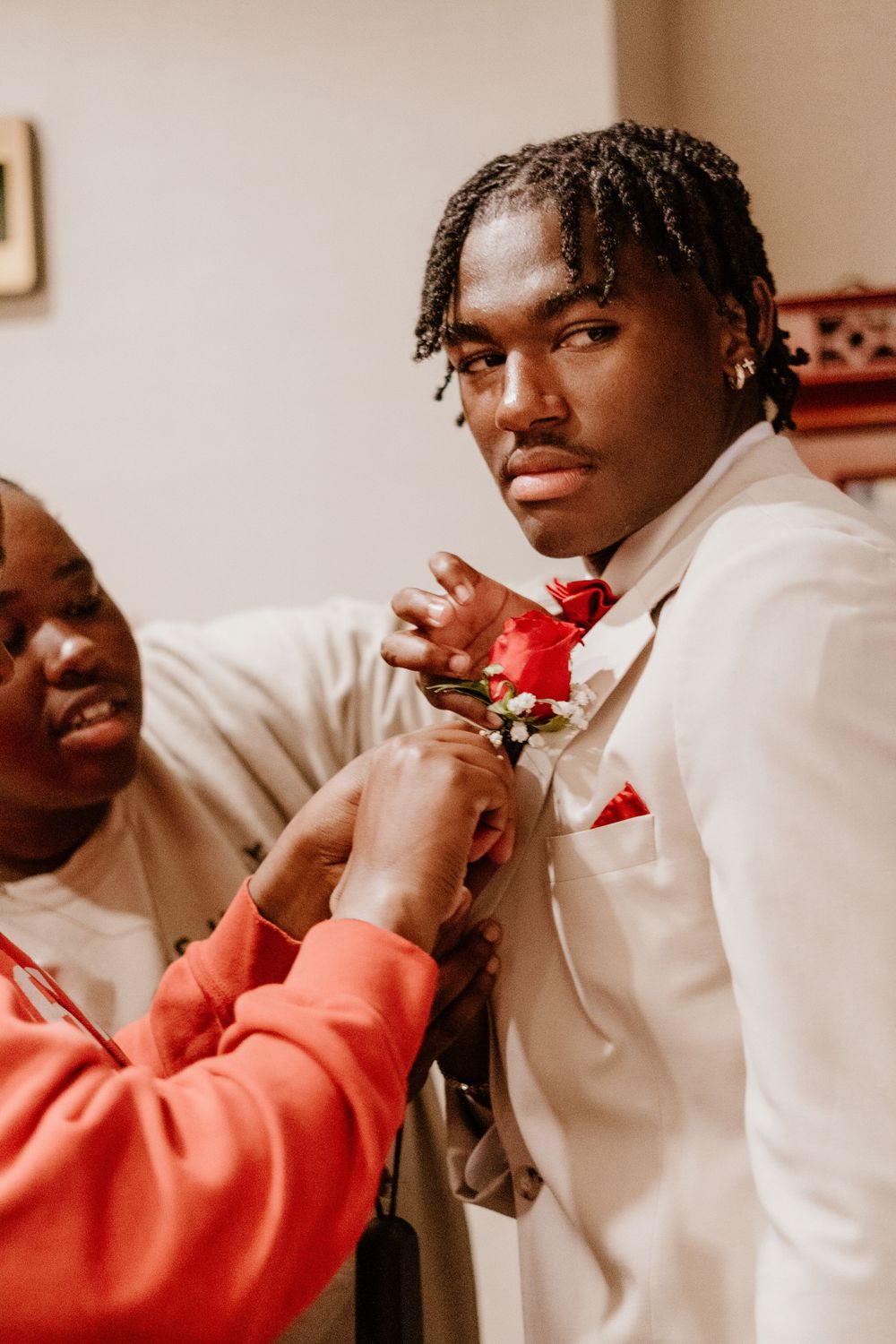 Person in white suit getting boutonniere pinned on by someone in coral colored clothing.